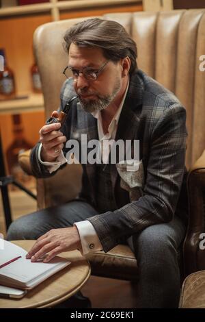 Reifen Geschäftsmann nachdenklich puffing Tabakpfeife im Tagebuch und Notizen zu machen. Älterer Mann im Restaurant arbeiten bis zum Abendessen serviert werden. Stockfoto