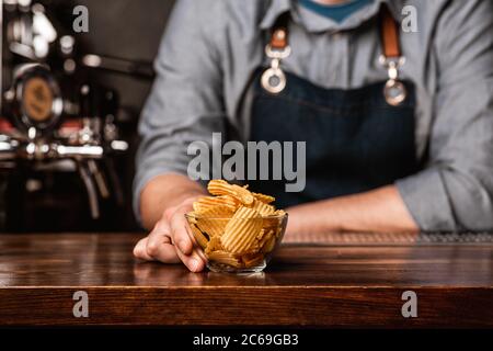 Snacks vom Barkeeper. Kerl in Schürze auf Holz braun Bar Zähler eine Glasplatte mit Chips Stockfoto