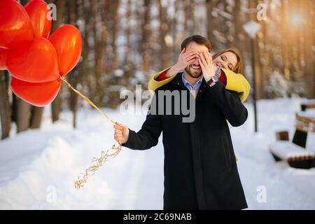 Junge kaukasischen Kerl stehen warten auf seine Freundin auf einem romantischen Date mit roten Luftballons. Schöne Dame schloss seine Augen hinter dem Rücken stehen. Gewinnen Stockfoto