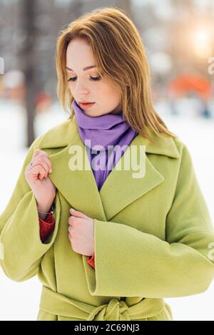 Portrait von hübschen kaukasischen Frau trägt grünen stilvollen Mantel und lila Schal. Kurzhaarige Dame bescheiden Blick nach unten. Verschneite Winter Straße in der BA Stockfoto