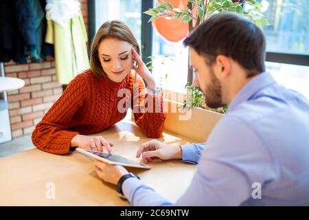 Portrait von schönen jungen kaukasischen Paar sitzt in modernen Café mit Tablet. Schöner Mann im Hemd und attratctive Dame in Bluse haben Mittagessen toge Stockfoto