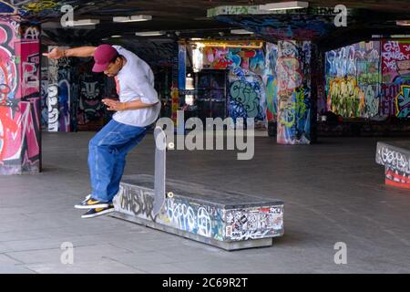 Mann in Baseballmütze, weißem T-Shirt und blauen Jeans, die mit Skateboard springen, im städtischen Graffiti-Skatepark auf der Londoner Southbank Stockfoto