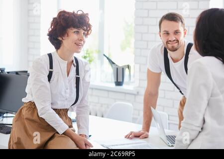 Schöne lächelnde Frau schaut Seite an Trainer, aufmerksam zu sprechen hören, Kollegen um sie herum, sitzen auf Tisch mit Computern, Papiere Stockfoto