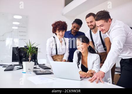 Junge und begeisterte Menschen im Büro, stehen in der Nähe asiatische Frau sitzen mit Laptop. Besprechen Sie das neue Projekt mit einem lapchenden Blick auf den Bildschirm des Laptops Stockfoto