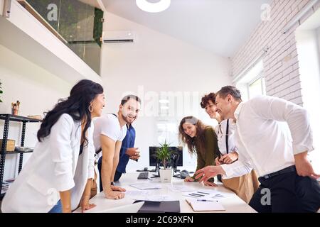 Multiethnische Gruppe von Menschen stehen um den Tisch herum und genießen Treffen, Lächeln und Lachen. Stehen Sie sich im weißen Büro gegenüber Stockfoto