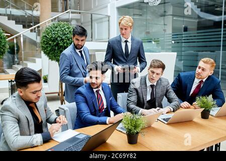 Internationale Gruppe von Geschäftsleuten, die auf dem Tisch sitzen, jeder schaut auf den Mann, der am Rand mit Laptop sitzt. Stockfoto