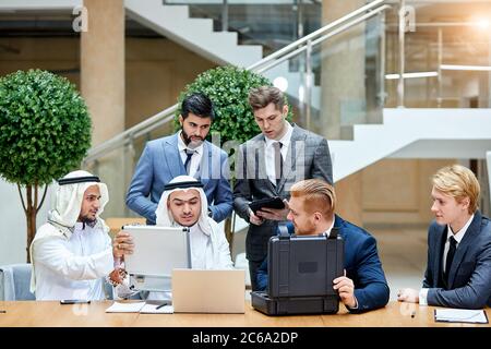 Kaukasische Geschäftsleute in Anzügen zeigen neue Technologie in schwarzem Gehäuse. Scheich zeigen Geld für Tauschhandel. Internationale Tagung im Business Center Stockfoto