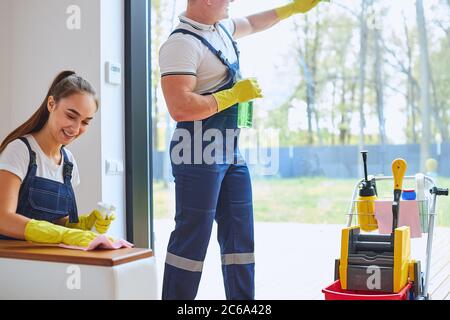 Seitenansicht auf aktive Arbeit von zwei kaukasischen Hausmeister in Uniform für die Reinigung gekleidet, Mann sauberes Glas Fenster, lachende Frau wischen Staub vom Sofa. Whij Stockfoto