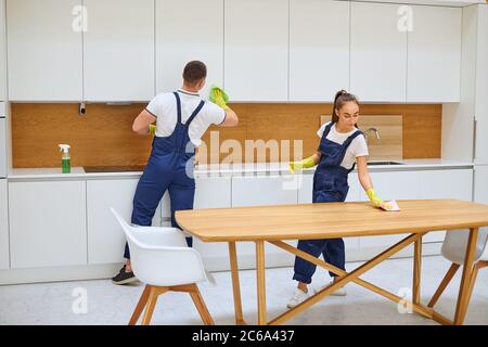 Junge kaukasische Reiniger in Uniform Reinigung Küche zusammen, wischen Staub von Holztisch. Weißer Innenraum Stockfoto