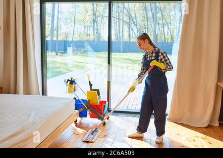 Freude an der Reinigung. Junge schöne Hausmeister Reiniger in blauer Uniform mit Reinigungsausrüstung stehen wischen den Boden im Schlafzimmer. Großes Panorama Stockfoto