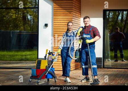 Zwei kaukasische Hausmeister, Putzfrauen stehen nach erfolgreicher Arbeit in einem großen Holzhaus mit Panoramafenstern posiert. Halten von Mopp, Reinigungsmittel Stockfoto