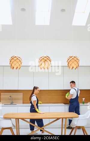 Junge kaukasische Reiniger in Uniform Reinigung Küche zusammen, wischen Staub von Holztisch. Weißer Innenraum mit Holzkegel Stockfoto