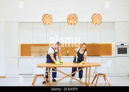 Junge Putzfrauen wischen Küchentisch, weiß europäischen Innenraum der Küche. Zwei Hausmeister tragen blaue Arbeitsuniform, gelbe Gummihandschuhe. holzkäfige Stockfoto