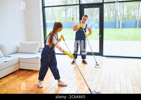 Junge schöne Mann und Frau in blauer Uniform halten Mops für das Waschen Wohnzimmer, effektive Arbeit, sprechen während des Wischbodens, arbeiten Stockfoto