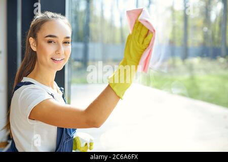 Vorsichtig junge Reiniger in gelben Gummihandschuhe Reinigung Fenster mit Spray und Lappen. Reinigungskonzept. Hausmeisterin Dame Blick auf Kamera Stockfoto