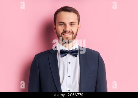 Junge schöne Mann Blick auf die Kamera mit Lächeln auf Gesicht, natürlichen Ausdruck. Lacht selbstbewussten Verlobten vor der Hochzeit. Rosa Hintergrund Stockfoto