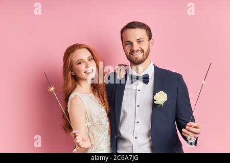 Kaukasisches Paar zelebriert ihre Ehe nach einem Jahr. Rothaarige Frau in weißem Kleid lächelnd, halten Wunderkerzen in den Händen. Mann in Smoking in der Liebe mit w Stockfoto