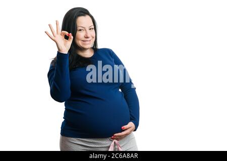 Lächelnde Frau hält Hand auf schwangeren Bauch machen okay Geste mit Fingern isoliert auf weißem Hintergrund Stockfoto