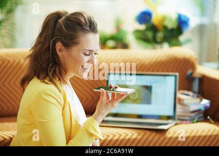 Lächelnd stilvolle 40 Jahre alte Hausfrau in gelber Jacke mit Laptop und Smartphone online im modernen Wohnzimmer an sonnigen Tagen studieren. Stockfoto