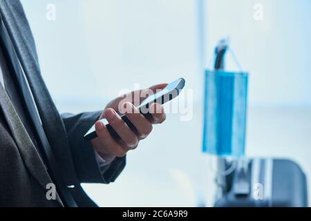 Nahaufnahme eines Geschäftsmannes, in einem grauen Anzug, sitzend in der Wartehalle eines Flughafens oder eines Bahnhofs, mit seinem Smartphone und einer chirurgischen Maske Hangi Stockfoto