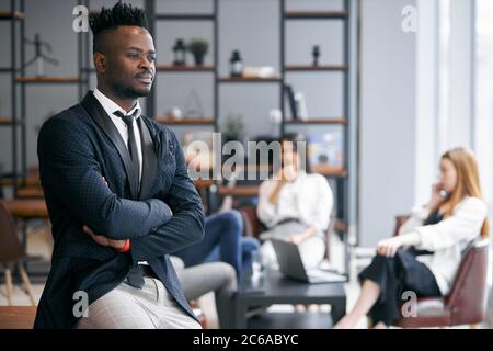 Gut aussehende afrikanische Mann trägt schwarzen Anzug Stand posiert in modernen Büro und Blick auf die Kamera. Hintergrund der Kollegen Stockfoto