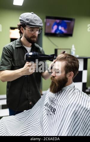 Sieht schon gut aus. Nahaufnahme der Seitenansicht des jungen bärtigen Mannes, der vom Friseur mit Haartrockner im Friseurladen gepflegt wird Stockfoto