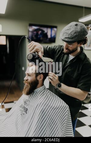 Sieht schon gut aus. Nahaufnahme der Seitenansicht des jungen bärtigen Mannes, der vom Friseur mit Haartrockner im Friseurladen gepflegt wird Stockfoto