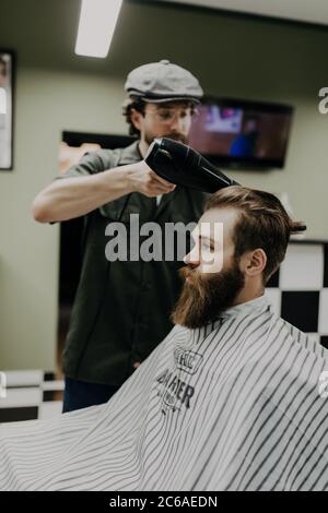 Sieht schon gut aus. Nahaufnahme der Seitenansicht des jungen bärtigen Mannes, der vom Friseur mit Haartrockner im Friseurladen gepflegt wird Stockfoto