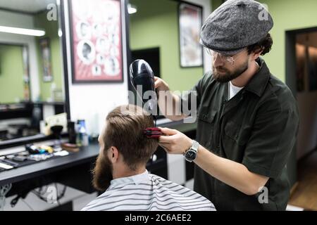 Sieht schon gut aus. Nahaufnahme der Seitenansicht des jungen bärtigen Mannes, der vom Friseur mit Haartrockner im Friseurladen gepflegt wird Stockfoto