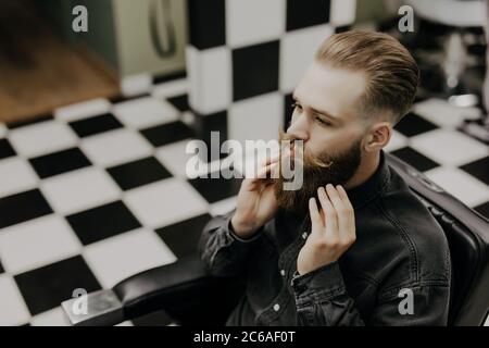 Hübscher junger bärtiger Mann auf sein Spiegelbild in den Spiegel schauen und halten die Hand in das Haar beim Sitzen im Stuhl bei Barbershop Stockfoto