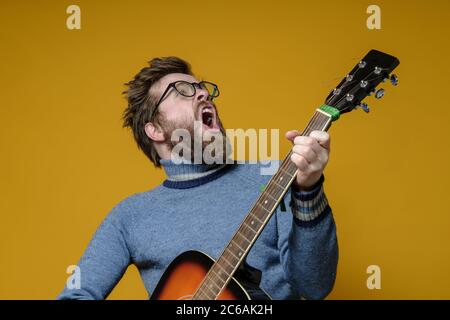 Hipster in einem alten Pullover spielt eine akustische Gitarre und singt laut, auf gelbem Hintergrund. Hobbys, Lifestyle. Stockfoto