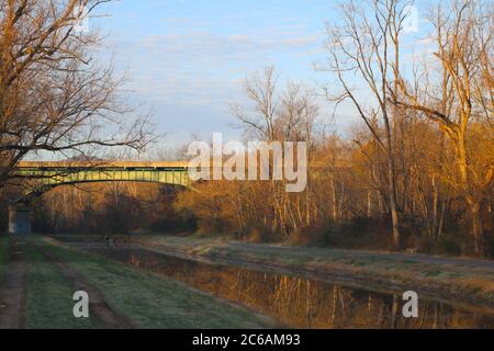 Eine hohe Stahlstraßenbrücke über einen ländlichen Kanal mit Spiegelung des Sonnenaufgangs und Wald Stockfoto