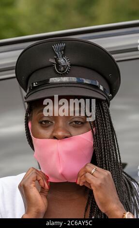 England, Großbritannien. 2020. Porträt einer Frau Chauffeur mit Uniform Kappe und geflochtenen Haaren Anpassung ihrer rosa gefärbten Gesichtsmask während der Covid-19 Ausbruch Stockfoto