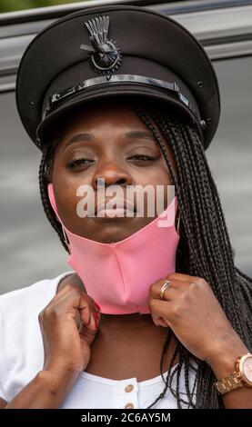 England, Großbritannien. 2020. Porträt einer Frau Chauffeur mit Uniform Kappe und geflochtenen Haaren Anpassung ihrer rosa gefärbten Gesichtsmask während der Covid-19 Ausbruch Stockfoto