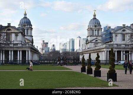 Zwei der gewölbten Gebäude des Old Royal Naval College mit Blick auf Canary Wharf. Ein Hundespaziergänger und zwei Touristen laufen auf dem Gelände. Stockfoto