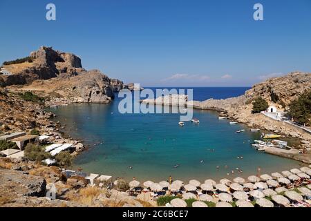 Die Lindian Akropolis erhebt sich über St. Paul's Bay, einer Bucht, die von Sonnenschirmen gesäumt ist, an einem sonnigen Tag in Lindos auf Rhodos, Dodekanes, griechischen Inseln, Griechenland, EU Stockfoto