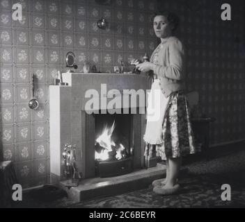 1950er Jahre, historisch, eine Hausfrau, die eine Schürze über einem Kleid mit Blumenmuster trägt, eine Wolljacke und Hausschuhe, die in einem Zimmer vor einem brennenden offenen Kohlefeuer in einem Vorzimmer stehen und eine Uhr vom gefliesten Kaminsims in England, Großbritannien, reinigen. Stockfoto