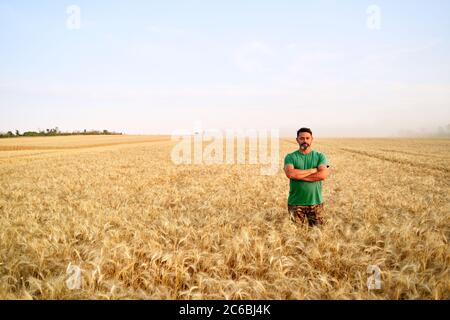 Glücklicher Bauer stolz im Weizenfeld mit gekreuzten Armen auf der Brust. Agronom trägt Corporate Uniform, Blick auf die Kamera auf Ackerland. Reich Stockfoto