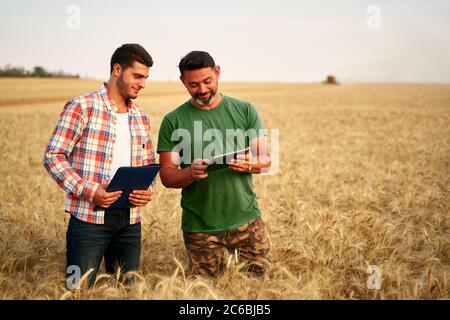 Zwei Bauern stehen im Weizenstoppelfeld, diskutieren Ernte, Ernte. Senior Agronom mit Touch Tablet pc lehrt junge Mitarbeiter. Innovative Technik Stockfoto