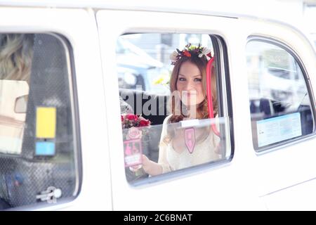 Zwanziger Rotschopf Boho Braut im Hochzeitskleid mit Girlanden in roten Ingwer Haar mit einem Blumenstrauß kommt zu ihrer Hochzeit in einem weißen Taxi. Stockfoto