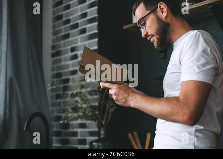 Junger Kaukasischer bärtiger Mann liest in der Küche ein Buch. Freizeit zu Hause. Stockfoto
