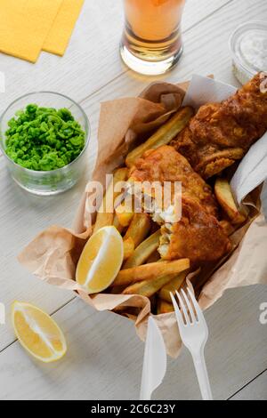 Fisch und Chips in Papier eingewickelt. Pürierte grüne Erbsen und Zitronenscheiben Stockfoto