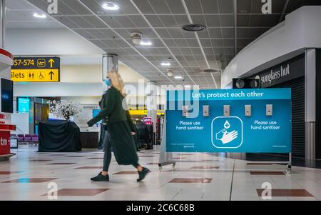 Eine Frau mit Gesichtsmaske geht während der weltweiten Coronavirus-Pandemie 2020, England, an einer Station für Händedesinfektionsmittel am Flughafen Gatwick vorbei Stockfoto