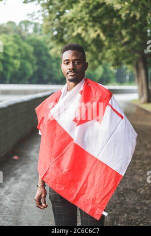 Schöner afroamerikanischer Mann mit kanadischer Flagge auf den Schultern, der ernsthaft auf die Kamera schaut und draußen steht. Stockfoto