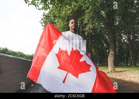 Schöner afroamerikanischer Mann mit kanadischer Flagge, der ernsthaft auf die Kamera schaut und draußen steht. Stockfoto