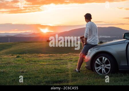 Junger Mann ruht während lange Solo Auto Reise sitzen auf seiner Luxus-Auto Motorhaube und genießen Sie den Sonnenuntergang Himmel Farben. Das Konzept „Reisen mit dem Auto“. Stockfoto
