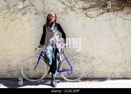 Junge Frau, die an sonnigen Tagen mit einem Vintage-Fahrrad an einer Stadtmauer angelehnt ist Stockfoto