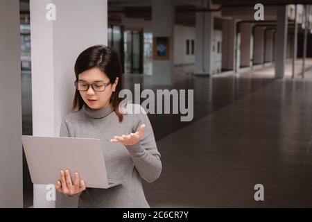 Frau im Büro hat Videogespräch. Junge positive Frau in Brille vor dem Laptop stehen und mit jemandem online sprechen. Stockfoto
