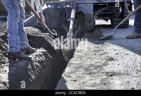 Nahaufnahme einer Trencher-Maschine in Bewegung Graben einen Graben in Schmutz mit Arbeitern schaufeln zur Seite Stockfoto