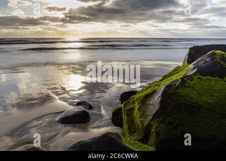 Die untergehende Sonne grast moosbedeckte Felsbrocken, während sich der Himmel auf dem nassen Sand in Carlsbad, CA, reflektiert. Stockfoto